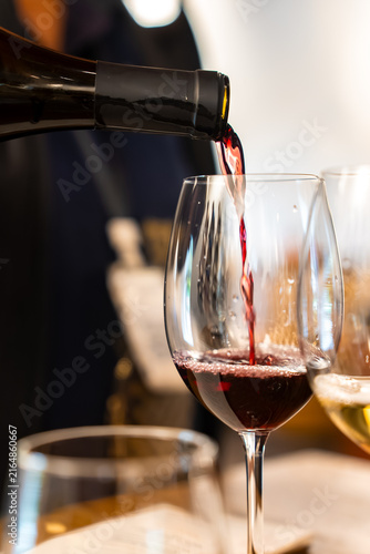 Foto Dark red wine pours smoothly from an open dark bottle neck into a stemmed clear glass during an indoor drink tasting session