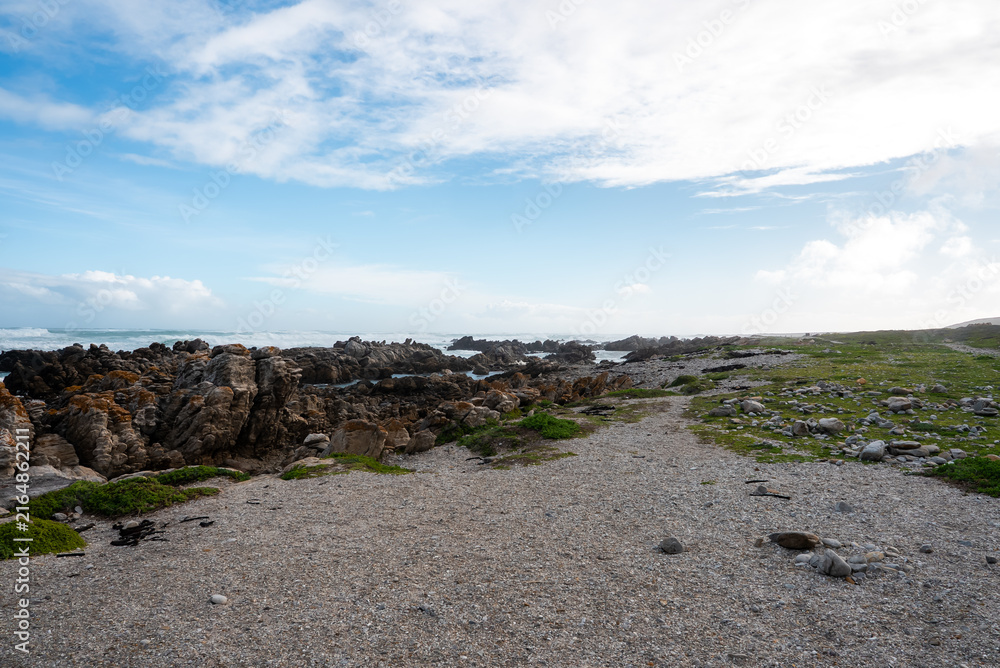 Obraz premium Gravel trail extends along the rugged rocky shoreline under a cloudy sky at Cape Agulhas in the Western Cape region of South Africa.