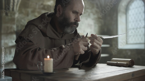 Fotografía Medieval monk sharpening a quill pen with a knife at a wooden table near a burni