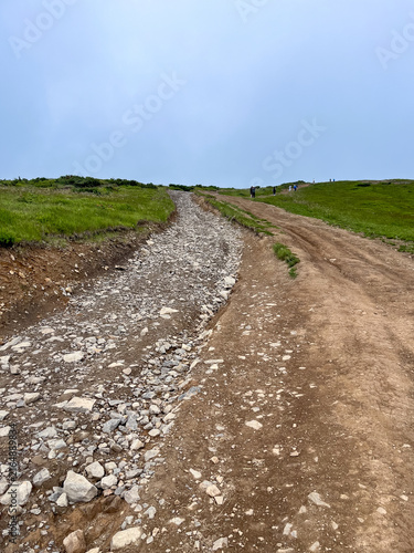 Cuadro en lienzo Hikers ascend a rocky and dirt path on a green grassy hill under a cloudy sky, representing journey and effort