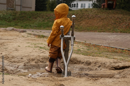 Obraz na plátne Toddler in a yellow raincoat and rubber boots playing in a muddy puddle on a playground, exploring water and sand with outdoor equipment