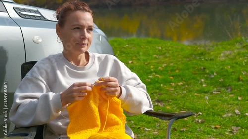 Woman knitting a while camping by a river. Middle-aged woman enjoying a relaxing outdoor hobby, knitting a warm yellow scarf while sitting in a camping chair next to her car on a autumn day by a river