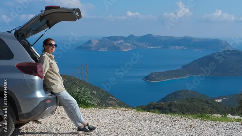 Tourist enjoying scenic view from car trunk. Serene female solo traveler leaning against open car trunk, enjoying breathtaking panoramic ocean landscape from scenic mountain viewpoint during road trip