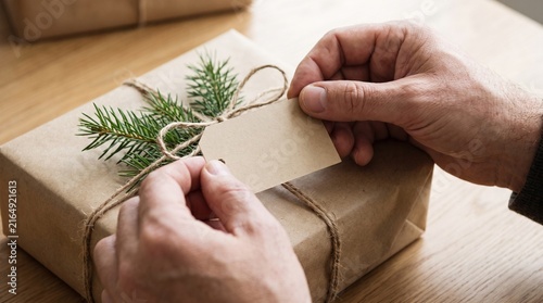 Hands tying twine around a kraft paper gift box decorated with a pine sprig and blank tag