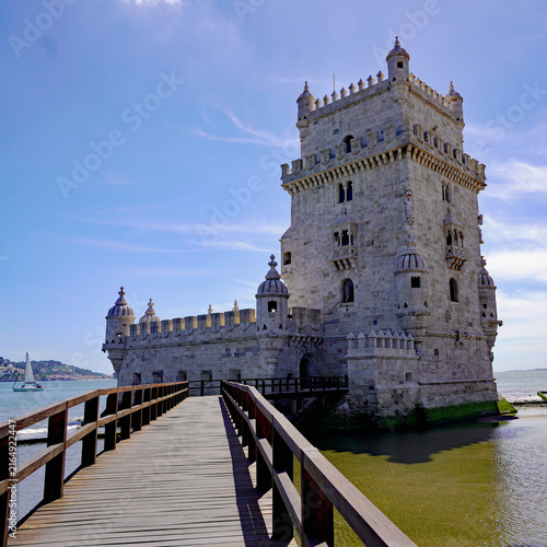A wooden walkway leads directly toward the iconic Manueline limestone fortress of Belém Tower rising from the waters of the Tagus River in Lisbon, Portugal