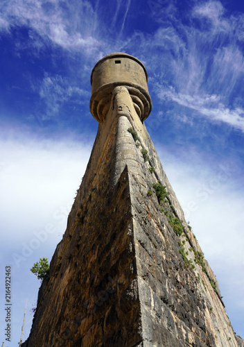 A dramatic low-angle perspective looks up the towering stone angle of the historic Cascais Citadel wall to a circular bartizan turret against a vivid blue sky in Cascais, Portugal