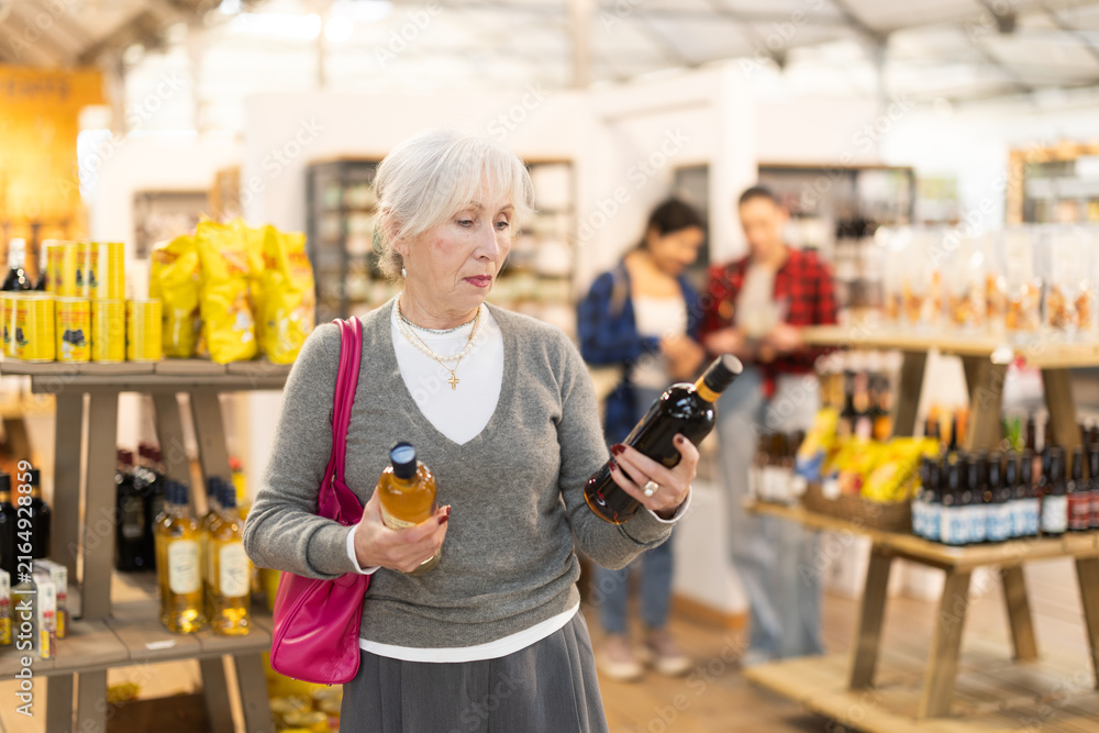 Fototapeta premium Elderly woman buyer chooses bottle of wine in grocery store