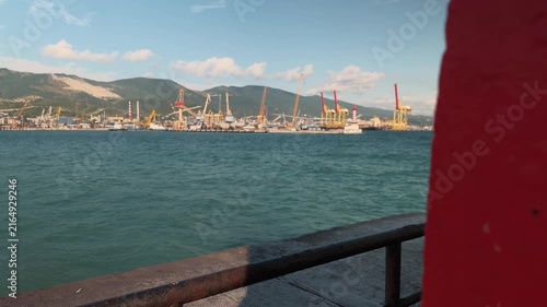 Peeking from behind a red wall at a large industrial seaport with yellow cargo cranes and shipping vessels in the sea with distant mountains under a blue sky for logistics and transport business