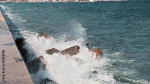 Ocean waves crashing into rusty concrete tetrapod breakwater blocks near stone pier with white foam and water splashes flying in the air on a sunny day authentic documentary coastal scenery