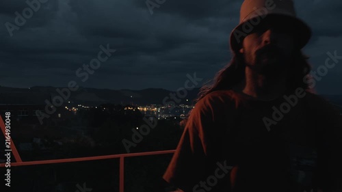 Solitary long-haired man in bucket hat stands on dark terrace leaning on railing looking at night city lights then turns and walks towards camera with face visible under dramatic cloudy dusk sky