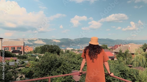 Unrecognizable long-haired man in bucket hat standing on private balcony admiring scenic panoramic view of coastal city seaport and mountains under blue cloudy sky during sunny day authentic rear view