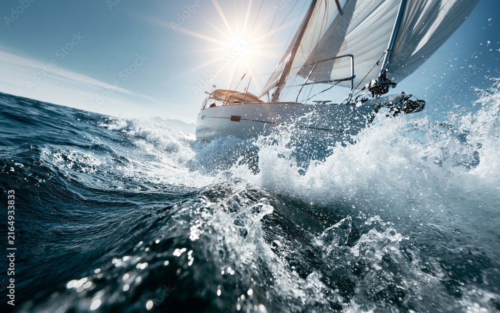 Obraz premium Sleek sailboat races through rough ocean waves. Water splashes high, creating white foam. Bright sun shines on blue sea. Dynamic yacht sailing fast during summer day.