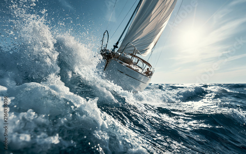 Sleek sailboat races through rough ocean waves. Water splashes high, creating white foam. Bright sun shines on blue sea. Dynamic yacht sailing fast during summer day.