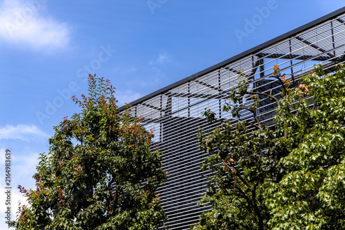 Photography Modern metal louvered building facade rises behind leafy green trees against a b