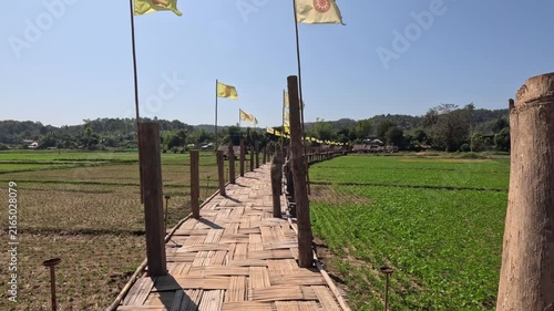 Wide shot of a wooden bamboo bridge with yellow flags on a clear day in northern Thailand, surrounded by rice fields, hills, and mountains near a Buddhist temple.