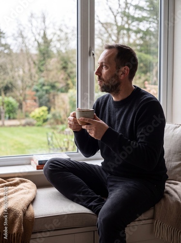 Pensive man sitting by a window at home holding a warm ceramic cup of coffee while looking thoughtfully at the peaceful garden outside