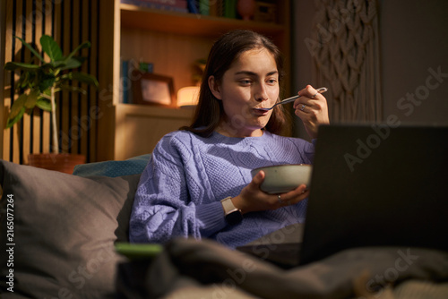 Foto Young adult Caucasian woman eating meal from bowl while watching laptop screen i