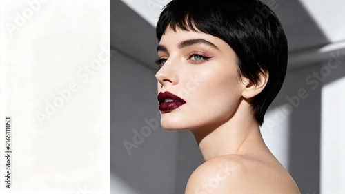 Profile of a woman with a chic pixie cut, bold burgundy lips, and flawless makeup, set against a minimalist architectural background.