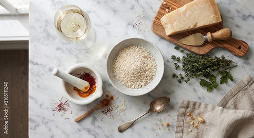 A top-down, artistic flat lay of a Milanese cooking setup featuring a glass of white wine, a small mortar and pestle with bright red saffron threads, a bowl of Arborio rice, and a block of Parmigiano 