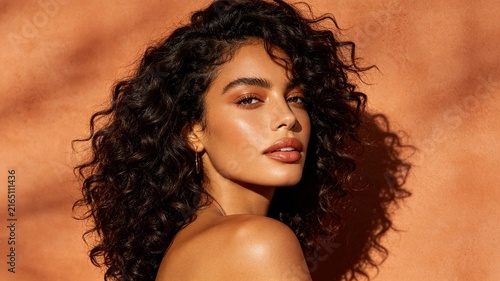 Portrait of a beautiful woman with voluminous dark curly hair and warm makeup, bathed in golden sunlight against a terracotta background.