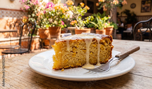 A rich slice of golden Pastel de Elote served on a white porcelain dish on a light wooden table, Moist, tender sweet corn cake texture with visible corn kernels, drizzled with sweet condensed milk and