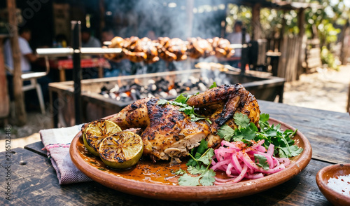 A rich half charcoal-grilled chicken with golden-brown charred skin served on a clay platter, Glistening citrus-marinated chicken served with charred lime wedges, pickled red onions, and fresh cilantr
