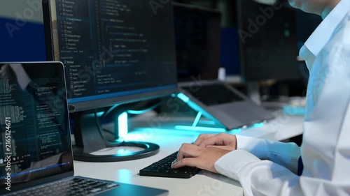 Software developer typing code on a keyboard and analyzing scripts on multiple screens in a dark, high-tech development office.