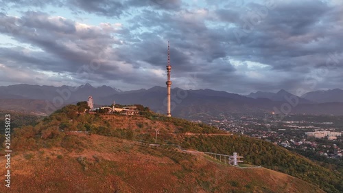 The famous TV tower on Kok Tobe Mountain in the Kazakh city of Almaty, set against a mountain range on a cloudy summer evening.