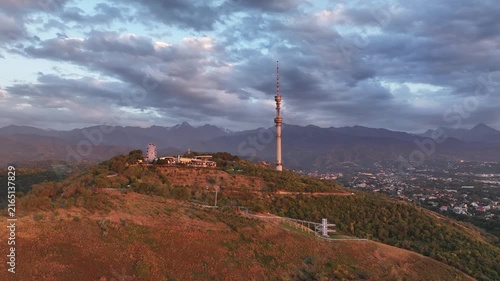 The famous TV tower on Kok Tobe Mountain in the Kazakh city of Almaty, set against a mountain range on a cloudy summer evening.
