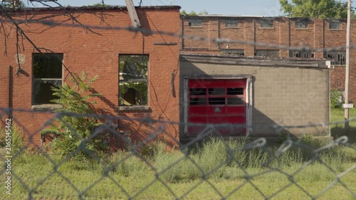 Slow zoom-in of an abandoned factory with a red garage door and broken windows. Chain-link fence. Old industrial building. Creepy.