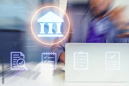 Working at a desk with documents and a bank icon on the computer screen in a busy office