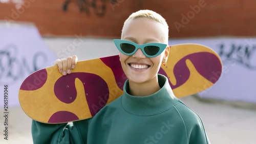 Smiling young woman skateboarder at a graffiti skatepark carrying a colorful skateboard on her shoulder wearing green sweatshirt and teal sunglasses for urban portrait background and banner design