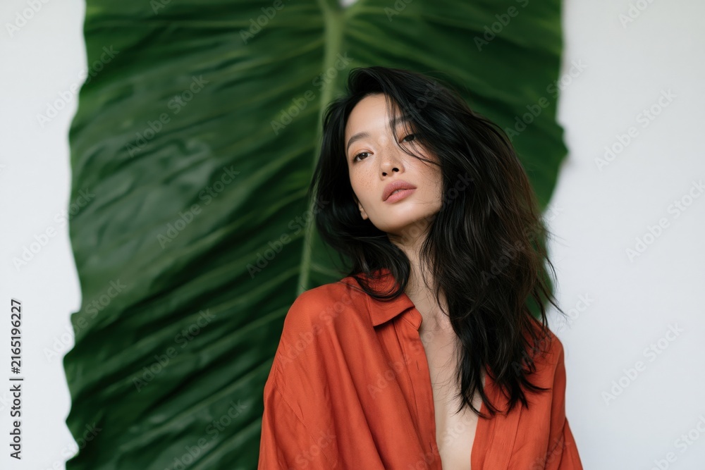 Fototapeta premium Elegant female portrait with oversized tropical leaf backdrop and terracotta shirt for beauty banner and fashion background
