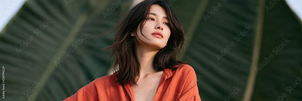 Fototapeta premium Portrait of a woman in an orange shirt against oversized tropical leaf background for fashion banner and editorial background design