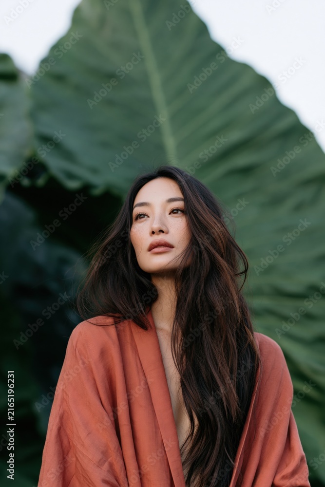 Fototapeta premium Vertical portrait of a woman draped in an orange robe against a giant tropical leaf background for editorial poster and wallpaper design