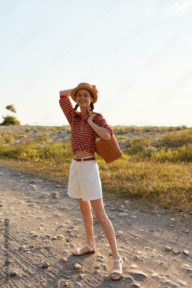 Fototapeta premium Young woman in a striped red top and white shorts poses outdoors under bright sunlight, smiling while carrying a woven bag in a casual summer lifestyle portrait, ideal for travel, fashion, or