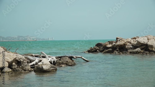 Picturesque sea coastal bay with turquoise water bordered by rocks. In distance of sea bay city appears on hill. Natural stone breakwaters near beach.
