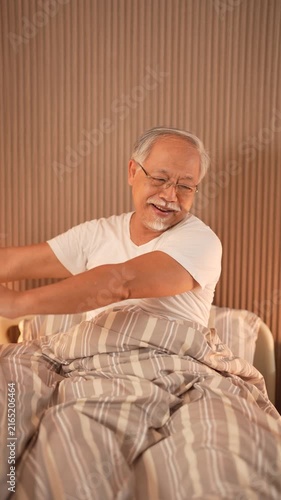 Senior man in glasses sits up in his hospital bed and stretches both arms wide as he wakes, then settles his hands on his lap. 4K elderly care b-roll filmed in a bright Thai nursing