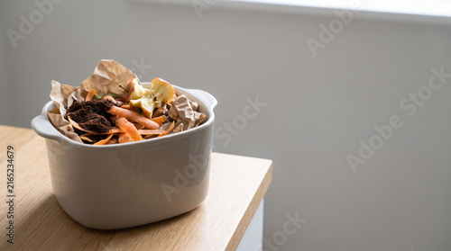 Vegetable scraps in compost caddy with coffee grounds and apple core on wooden table in bright room