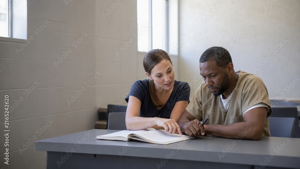 Obraz premium Female prison counselor assists male inmate with paperwork at a table in a correctional facility, bright windows providing natural light in the background