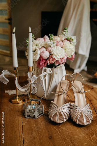 Wedding accessories on the wedding day for the bride, a white dress, decorated high-heeled shoes, a box with wedding rings, a bouquet of flowers and a candelabra with candles next to them on the table