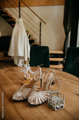 White bride's dress hanging near the stairs in a hotel room and beautiful shoes standing on a table next to a box with golden wedding rings on their wedding day