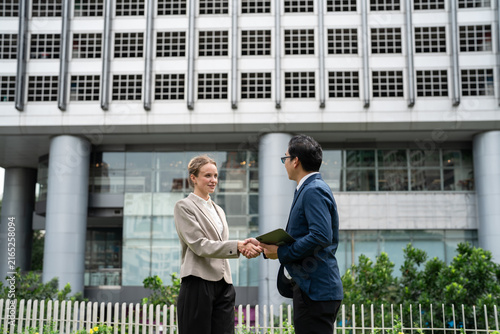 International business partners shaking hands outside modern office building, diverse businesswoman and businessman greeting agreement in corporate district