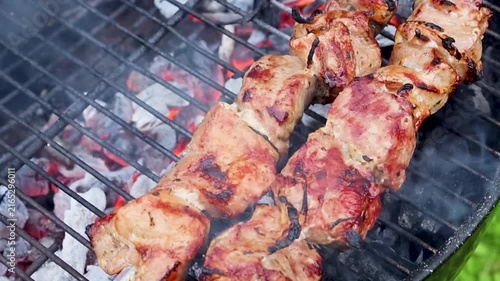 Marinated pieces of meat grilling on metal skewers over a hot charcoal grill in a summer garden with fresh green grass in the background. Close-up barbecue scene with smoky grilled meat