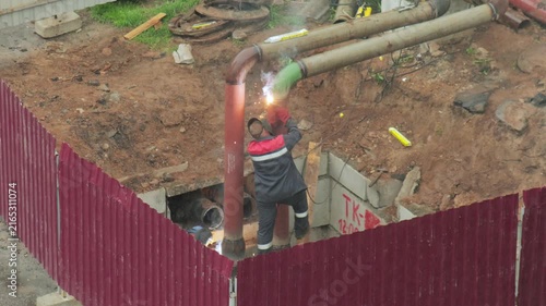 Welder works on pipe system in trench, bright sparks during joining process, industrial scene of construction worker performing duties with protective gear, professional at job site