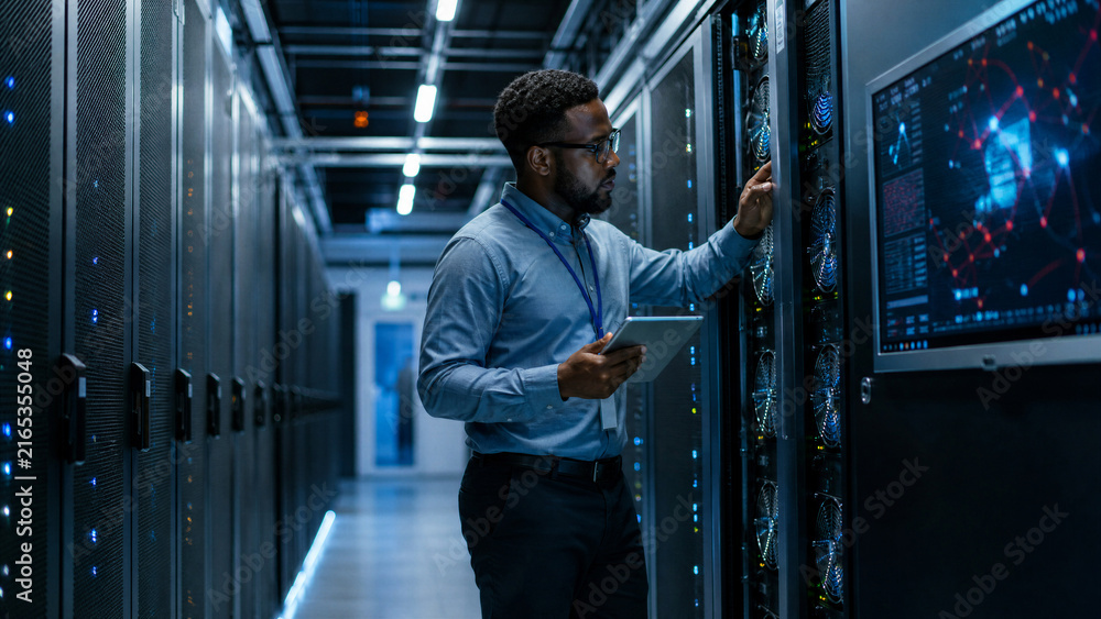 Fototapeta premium African American cybersecurity engineer holding a tablet while inspecting server racks in a high-tech data center