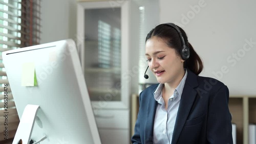 Professional call center agent providing customer support, smiling Asian operator wearing a headset with microphone and typing on a computer keyboard in a bright office