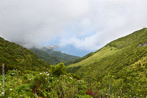 View from green hills to coast, glimpse through clouds of Ponta Delgada village and ocean