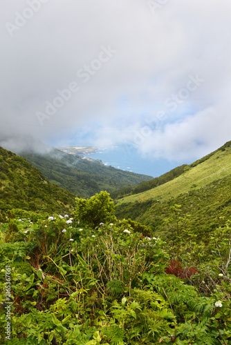 View from green hills to coast, glimpse through clouds of Ponta Delgada village and ocean
