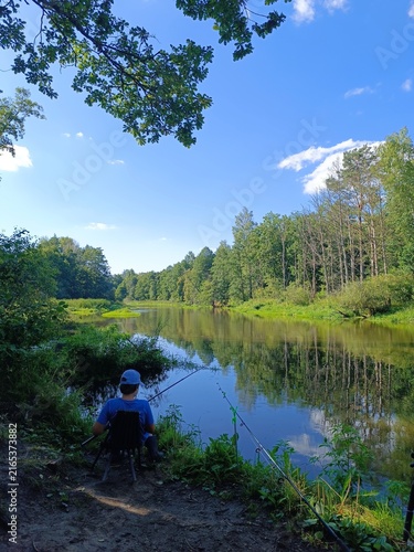 Person fishing by a river on a sunny summer day. Outdoor recreation and hobby in nature with green forest reflection